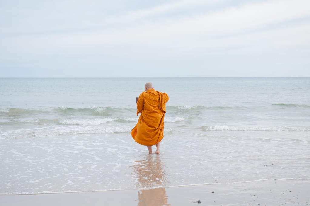 A monk in an orange robe standing barefoot at the edge of a calm beach, facing the ocean with gentle waves.