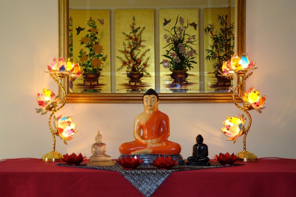A decorative altar displaying various Buddha statues, including a prominent orange figurine in a meditative pose, surrounded by lotus-shaped lamps and decorative flowers. The background features a mirror reflecting floral artwork.