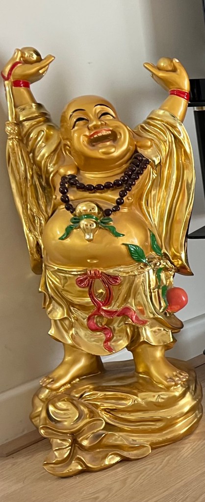 A large, gold-colored laughing Buddha figurine with a joyful expression, wearing colorful robes and adorned with beads. The Buddha is standing with arms raised, symbolizing prosperity and happiness.